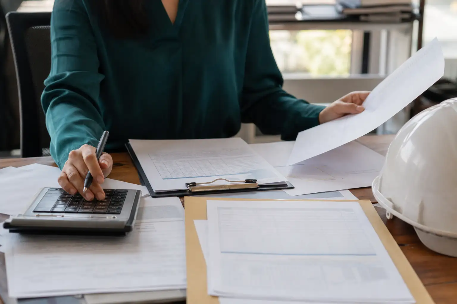 An accounts payable worker reviews a rental invoice, contract, and supporting paperwork at a desk with a hard hat and job-site documents nearby.