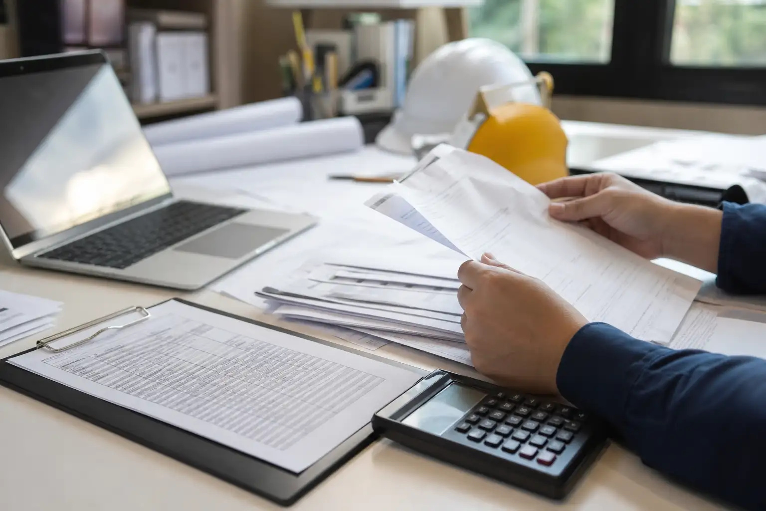 An office desk with rental invoices, a calculator, a laptop, and printed equipment delivery records being reviewed by a finance worker.