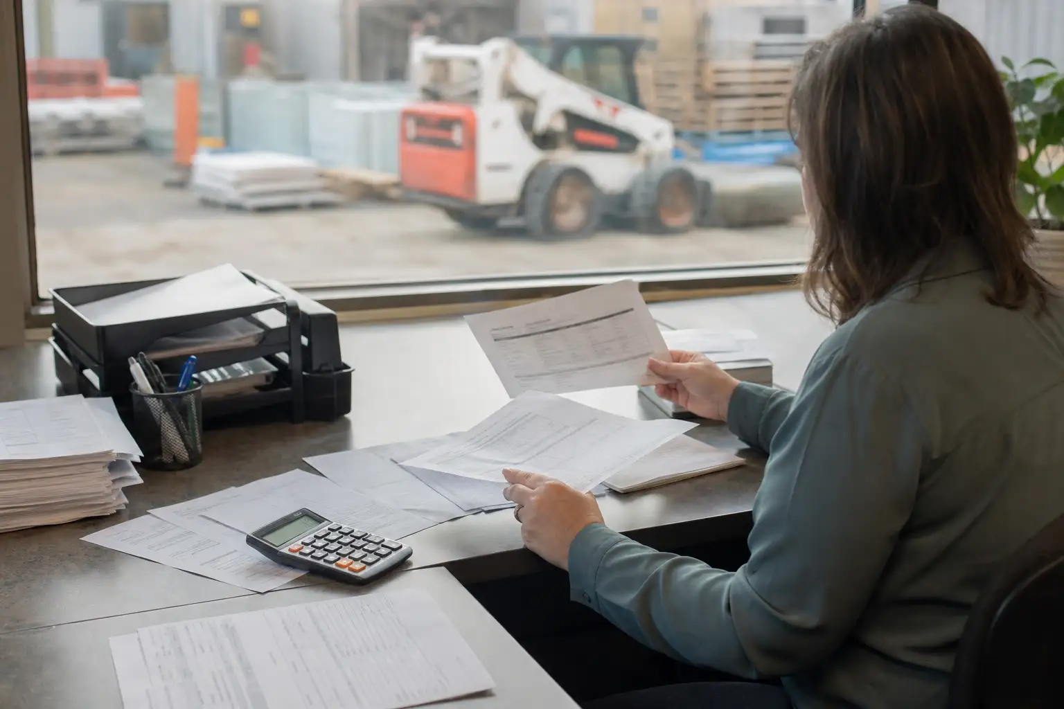 An AP employee reviewing rental paperwork, an off-rent confirmation, and a job coding sheet at a desk with construction equipment visible outside a window.