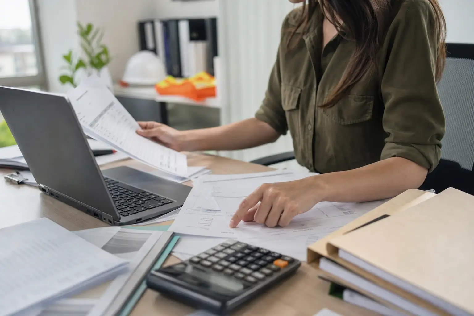 An AP specialist reviewing rental invoices, a laptop screen, printed paperwork, and project files on a desk in an office setting.
