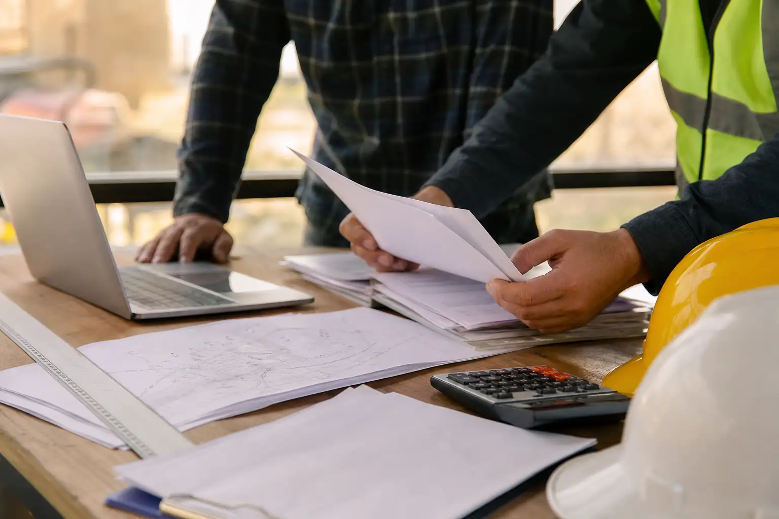 An office table with rental agreements, invoices, a laptop, and hard hats beside a site map