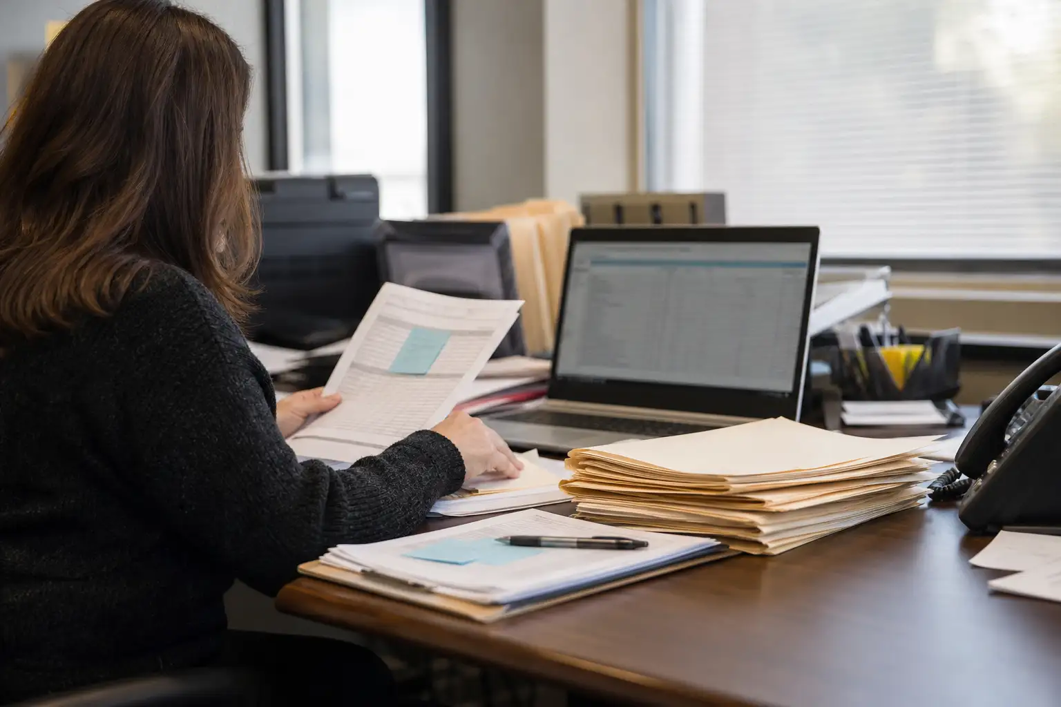 An AP specialist reviewing a stack of rental invoices, exception notes, and a laptop at a desk in a contractor office.